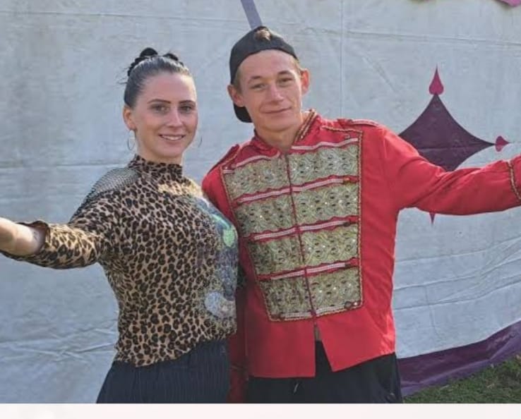 Two dancers pose arm in arm outdoors, woman in a leopard print top and man in a red decorative jacket, smiling at the camera.