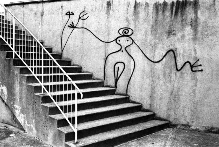 Black-and-white photo of graffiti on a concrete wall beside a stairwell: a long, abstract figure with a spiraled head and outstretched limbs drawn across the wall.