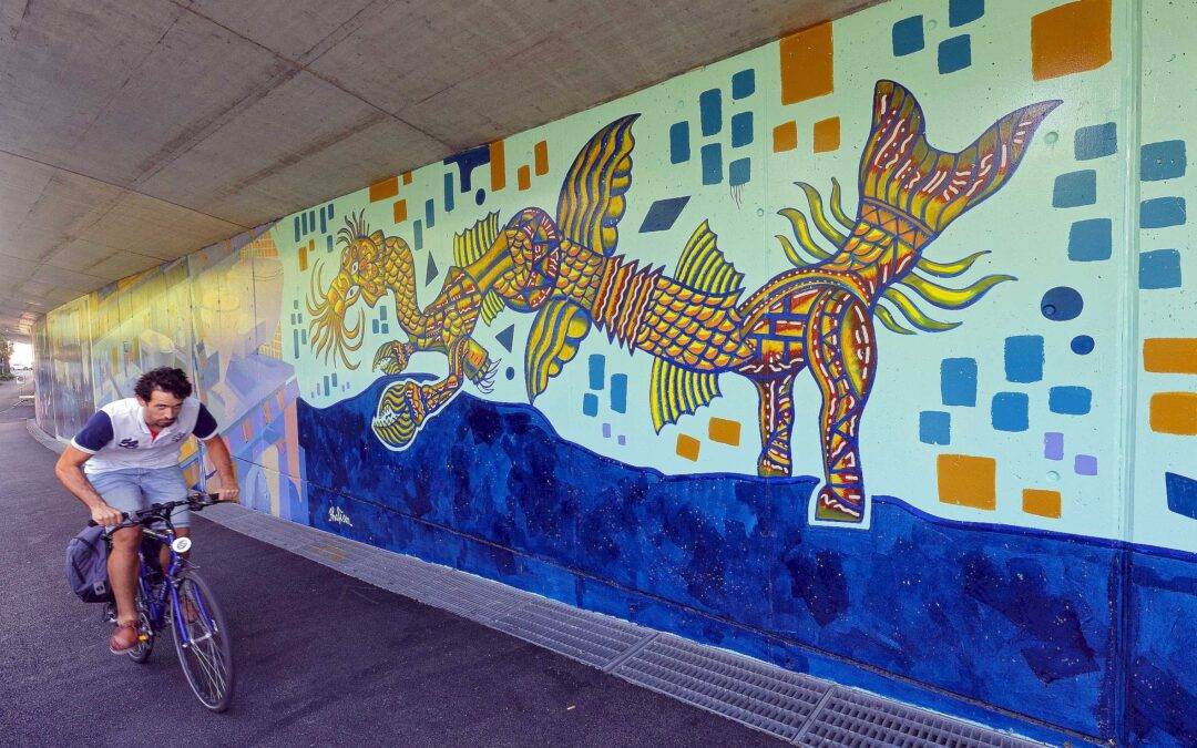 Man riding a blue bicycle under a tunnel wall covered with a bright mural of a fantastical sea-dragon creature and geometric shapes.