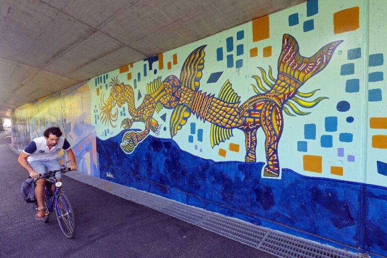 Man riding a blue bicycle under a tunnel wall covered with a bright mural of a fantastical sea-dragon creature and geometric shapes.