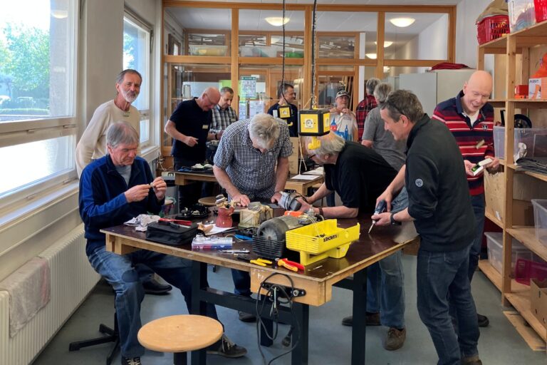 Group of men in a workshop at a long table repairing and assembling small devices; tools and parts scattered around.