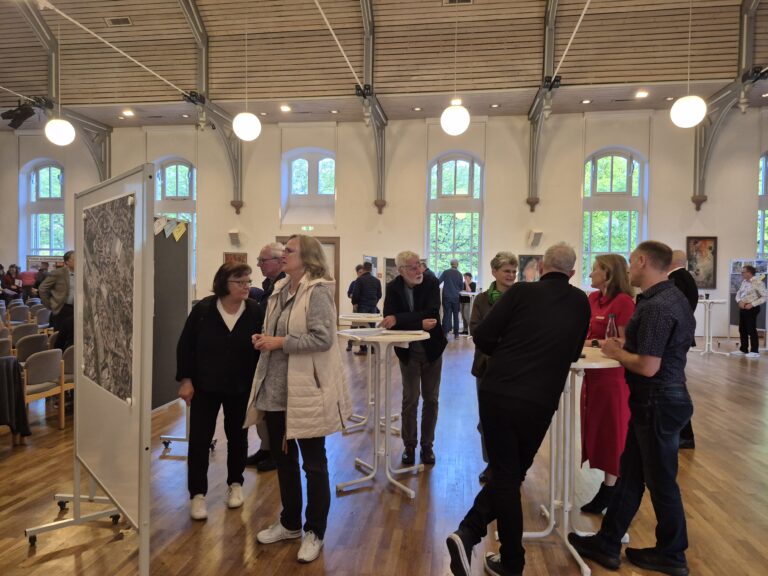 Group of people chatting around tall white tables in a bright gallery, with a large map display on wheels in the foreground and artwork on the walls behind them.