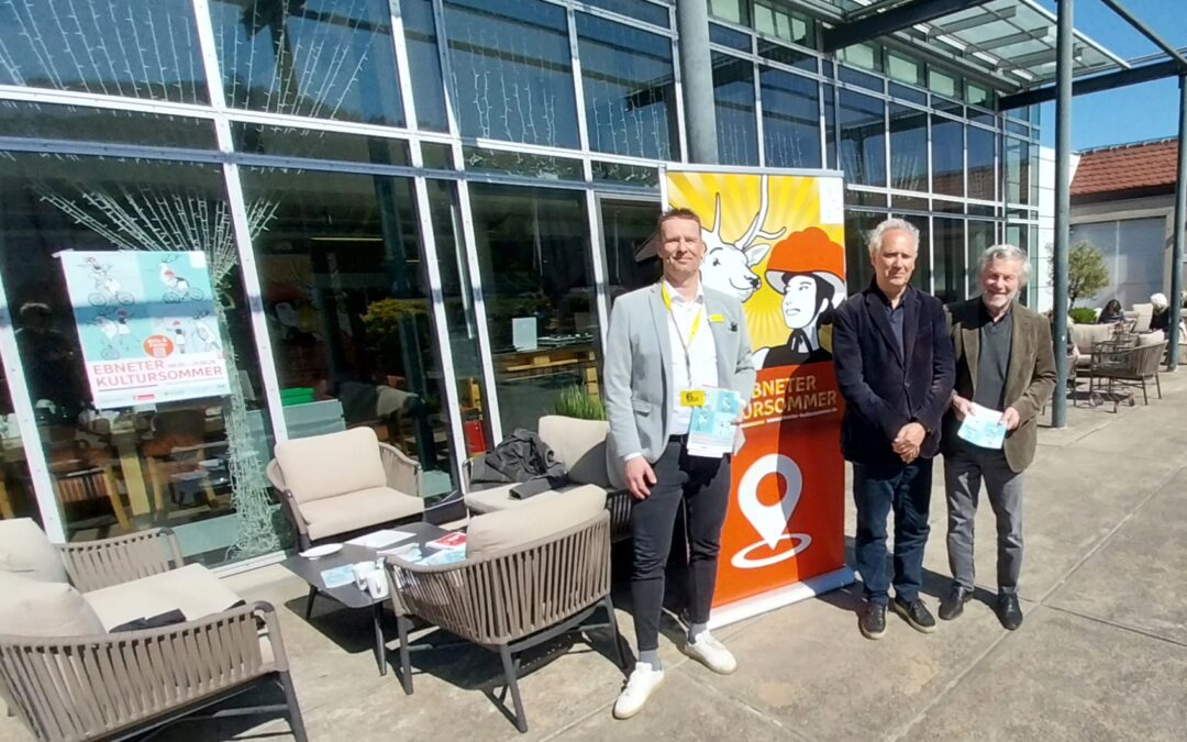 Three men stand together outside a glass-walled building beside a bright poster for a cultural event, posing for a photo.
