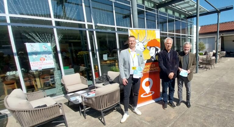 Three men stand together outside a glass-walled building beside a bright poster for a cultural event, posing for a photo.
