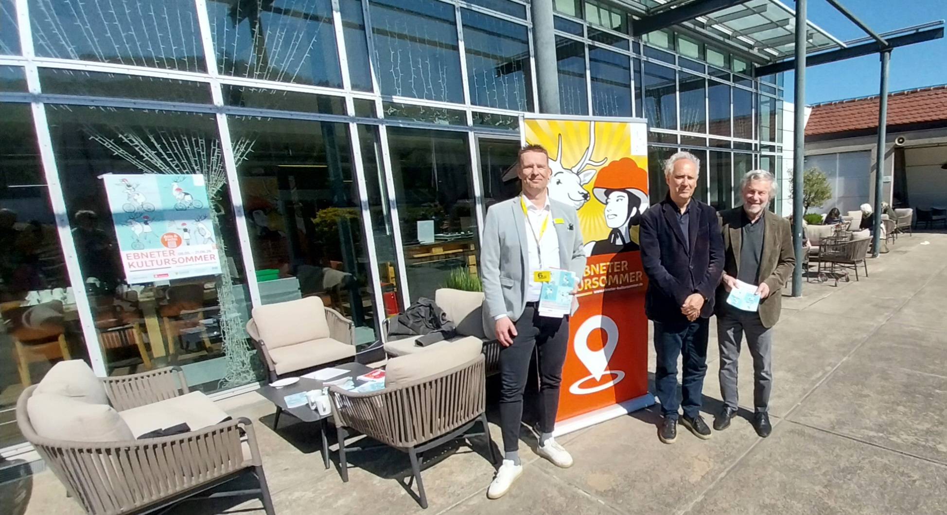 Three men stand together outside a glass-walled building beside a bright poster for a cultural event, posing for a photo.
