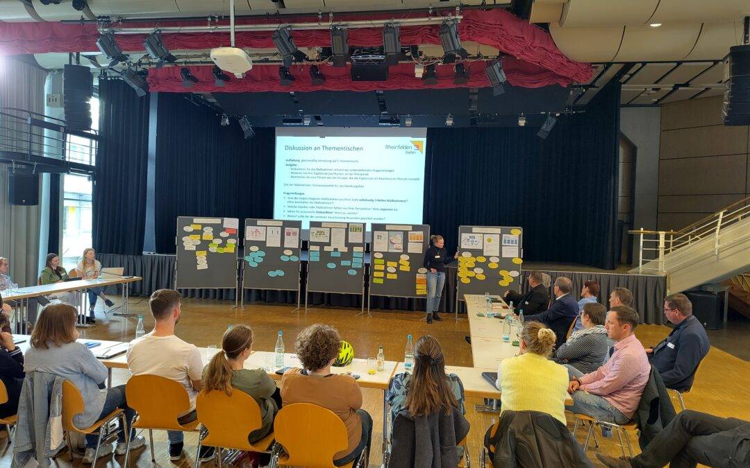 Conference hall with a stage, projector screen, and attendees seated at tables for a workshop or meeting. Large boards with notes line the front of the room.
