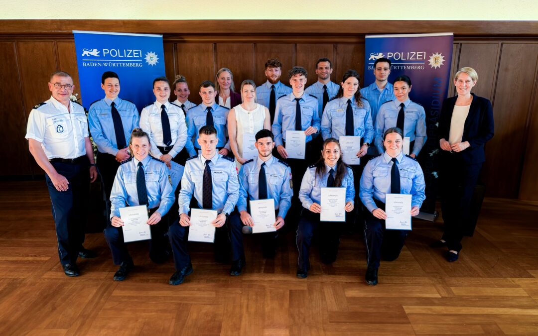 Group of police cadets in blue uniforms posing with certificates at a ceremony, Baden-Württemberg police banners in the background.