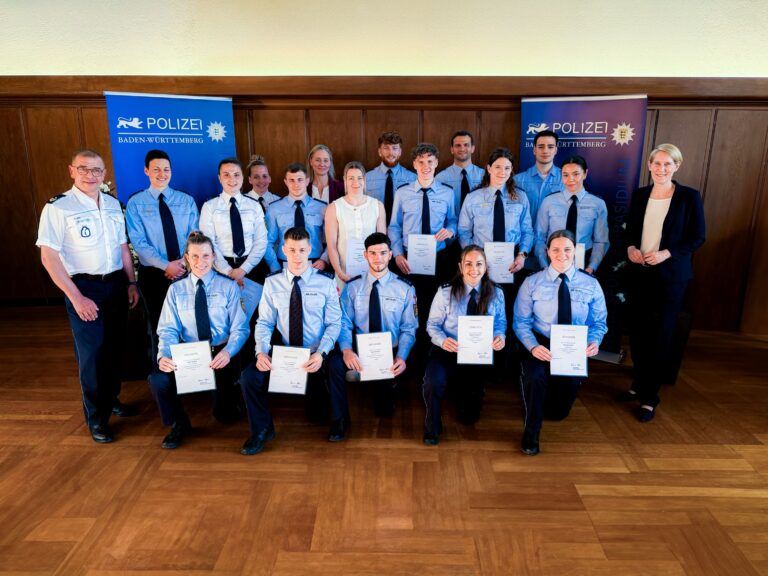 Group of police cadets in blue uniforms posing with certificates at a ceremony, Baden-Württemberg police banners in the background.