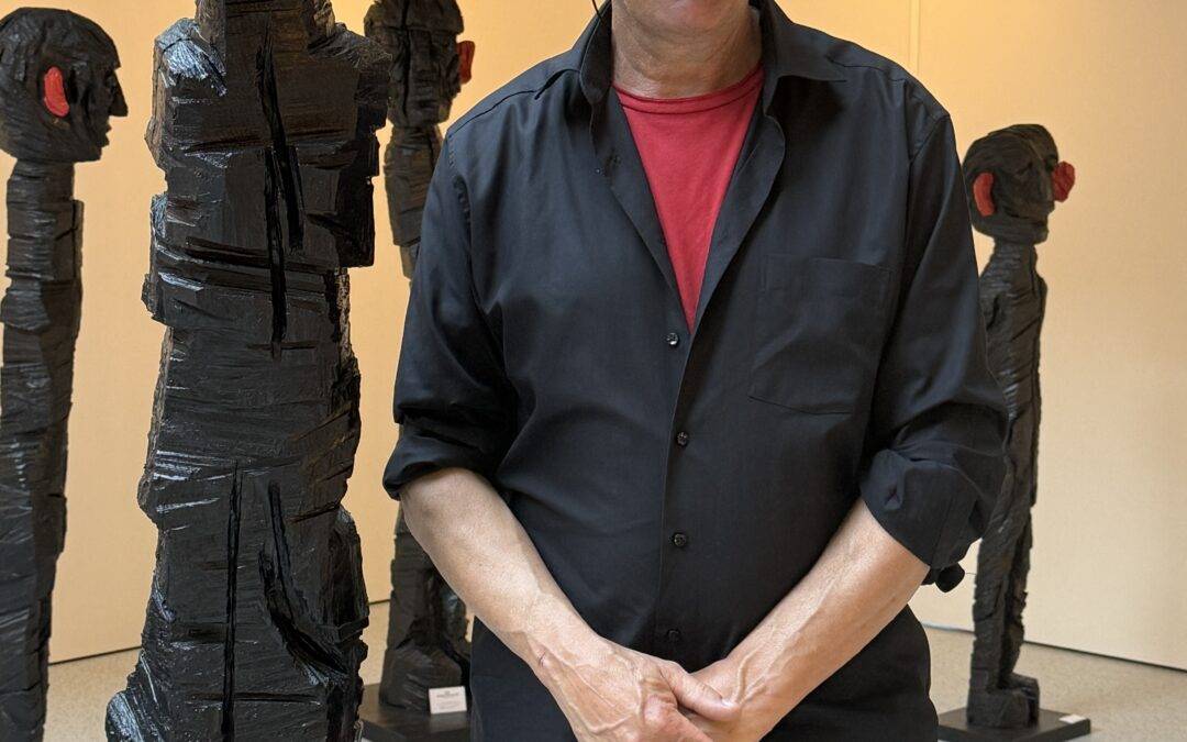 Man wearing black shirt over a red t-shirt, with glasses, posing in an art gallery beside tall, black wrapped sculptures with red accents on the ears/gears of the figures.