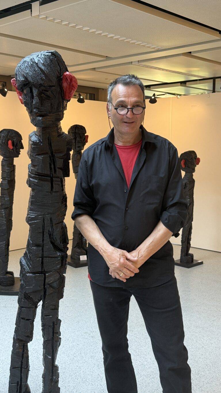 Man wearing black shirt over a red t-shirt, with glasses, posing in an art gallery beside tall, black wrapped sculptures with red accents on the ears/gears of the figures.