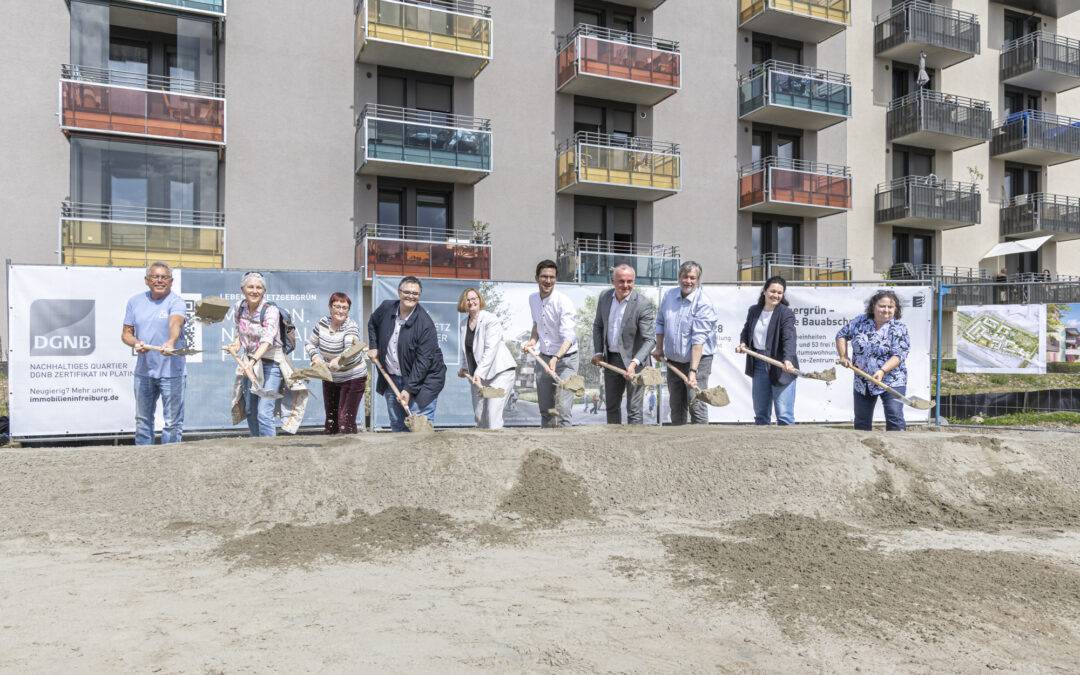 Group of people with shovels at a groundbreaking ceremony in front of a new residential building.