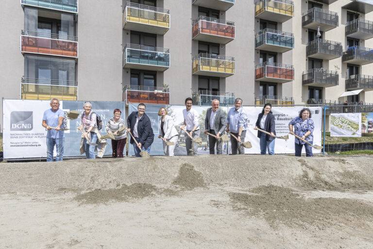Group of people with shovels at a groundbreaking ceremony in front of a new residential building.