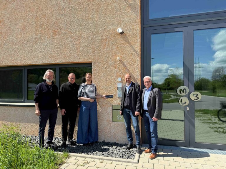 Five adults posing for a group photo outside a beige textured building with a large glass entrance door.