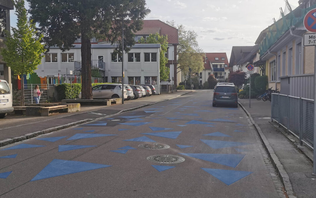 Urban street with blue triangular road markings, parked cars along both sides, and a no-parking sign on the right; residential buildings in the background.