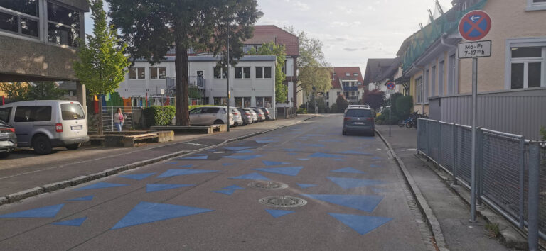 Urban street with blue triangular road markings, parked cars along both sides, and a no-parking sign on the right; residential buildings in the background.