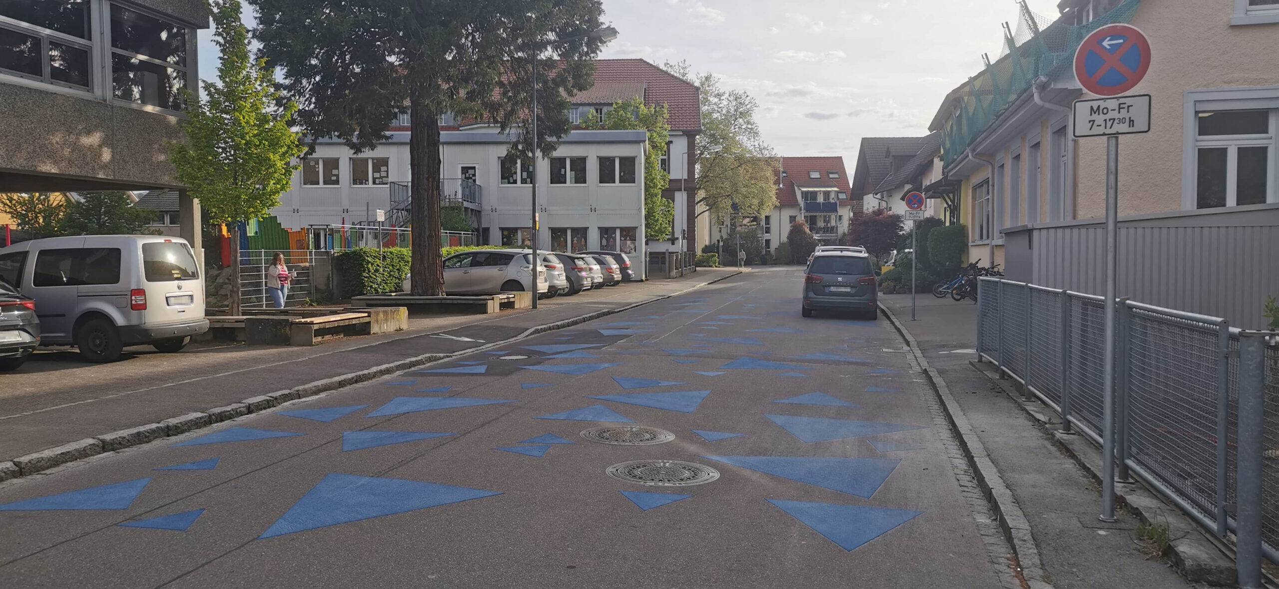 Urban street with blue triangular road markings, parked cars along both sides, and a no-parking sign on the right; residential buildings in the background.