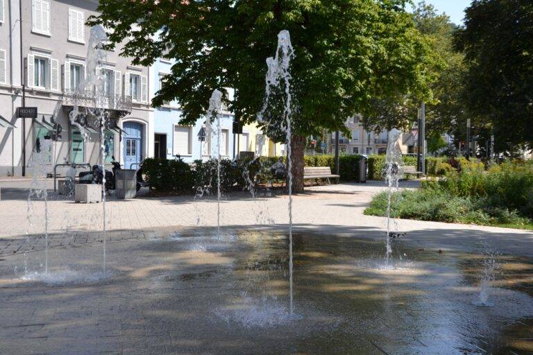 Public plaza with rising water jets in a shallow pool, tree-lined street and pastel buildings.
