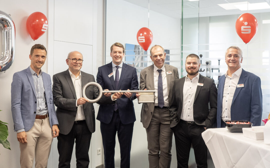 Six executives in business attire pose with a giant silver ceremonial key at a corporate event, in a modern office setting with red logo balloons nearby.