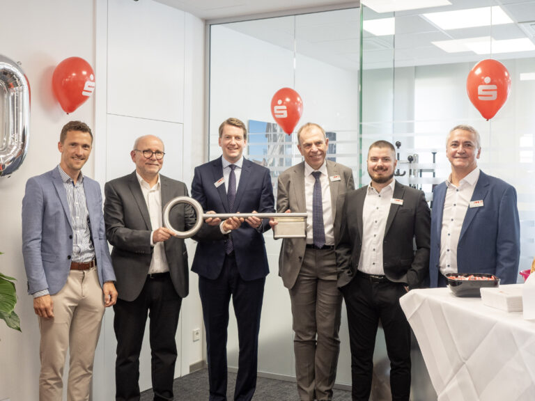 Six executives in business attire pose with a giant silver ceremonial key at a corporate event, in a modern office setting with red logo balloons nearby.