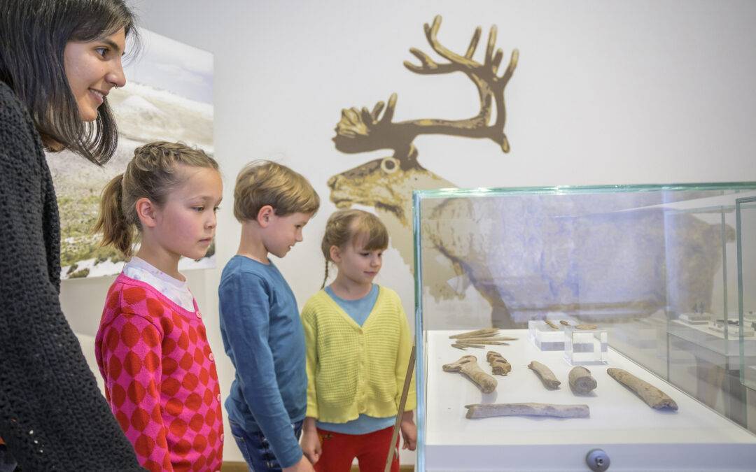 Woman and three children observe fossil bones in a glass display case at a museum, with a deer mural in the background.