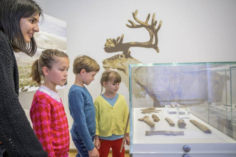 Woman and three children observe fossil bones in a glass display case at a museum, with a deer mural in the background.