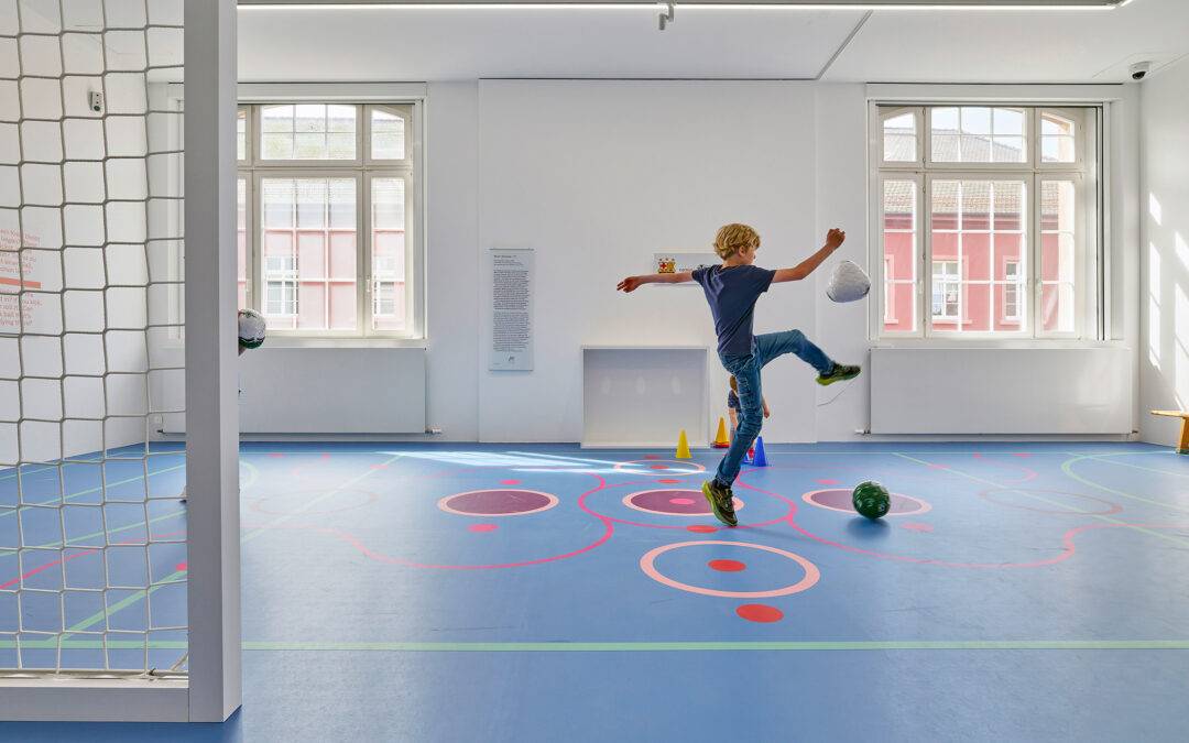 Boy kicking a soccer ball indoors on a blue gym floor with cones and large windows in the background.