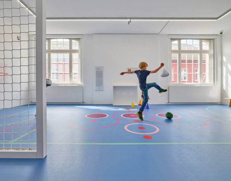 Boy kicking a soccer ball indoors on a blue gym floor with cones and large windows in the background.