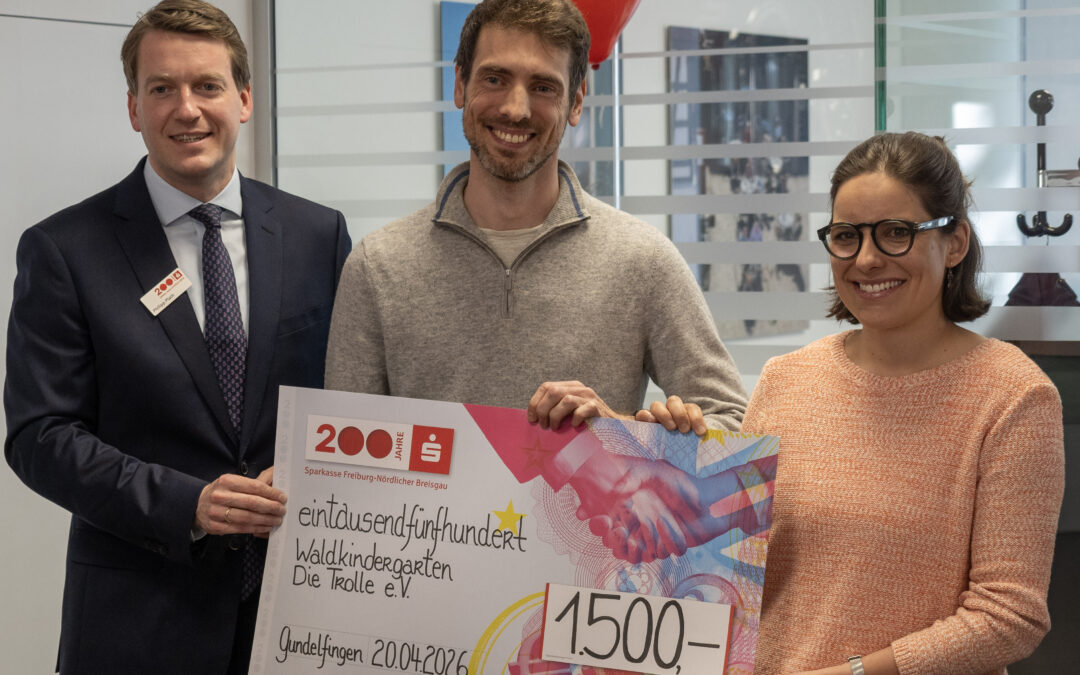 Three smiling people pose with a large colorful ceremonial cheque; a man in a dark suit stands on the left, a man in a gray sweater in the middle, and a woman with glasses on the right.