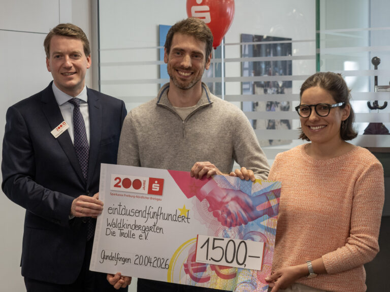 Three smiling people pose with a large colorful ceremonial cheque; a man in a dark suit stands on the left, a man in a gray sweater in the middle, and a woman with glasses on the right.