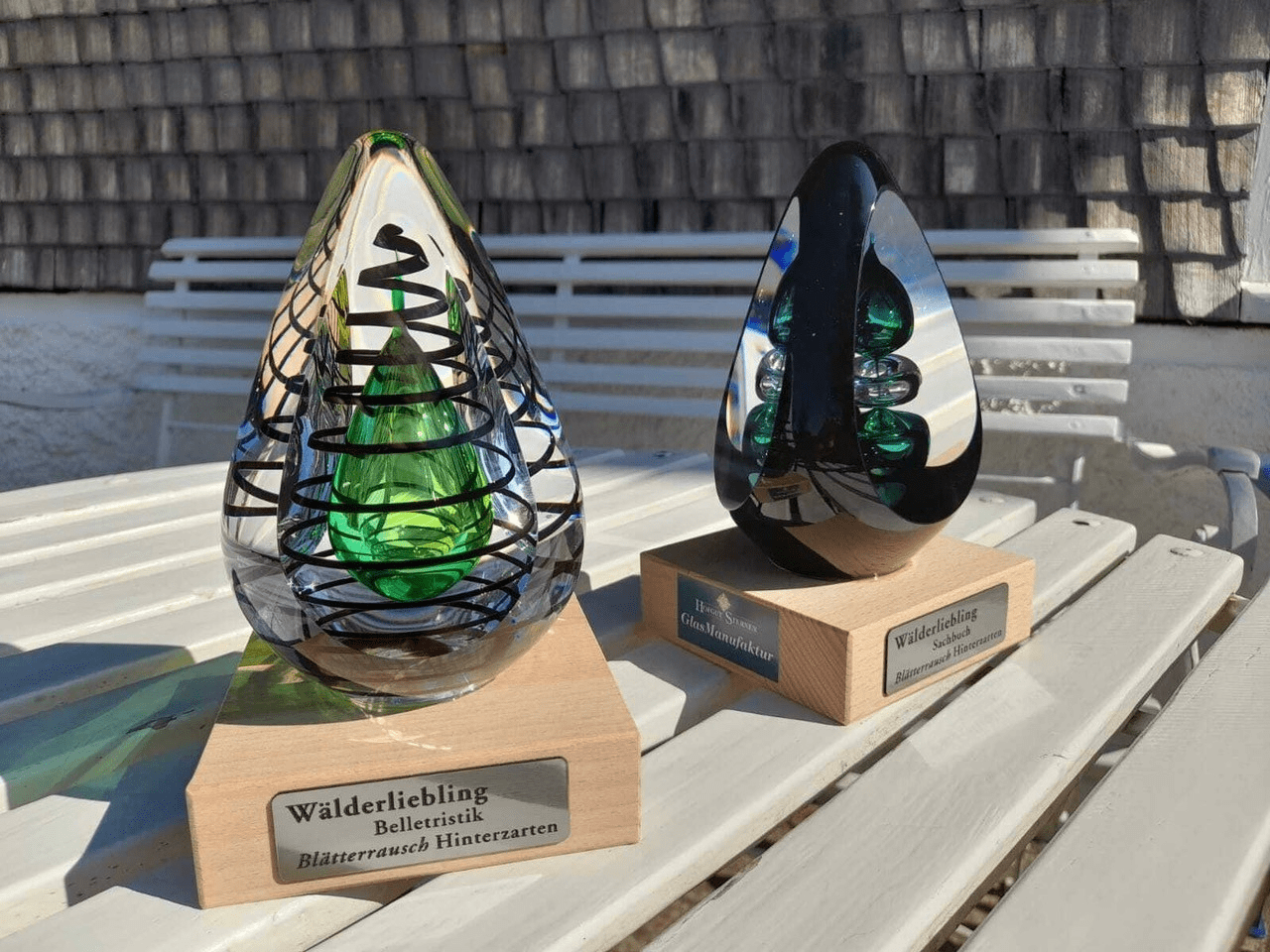 Two decorative glass teardrop sculptures on wooden bases displayed on a white bench, one green and clear with a black spiral design, the other dark with blue-green highlights.