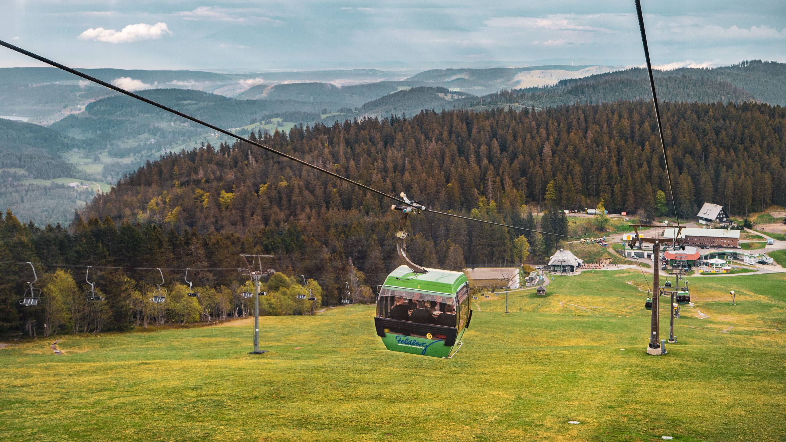 Green gondola cable car carrying passengers over a grassy hill with a forest and distant village below.