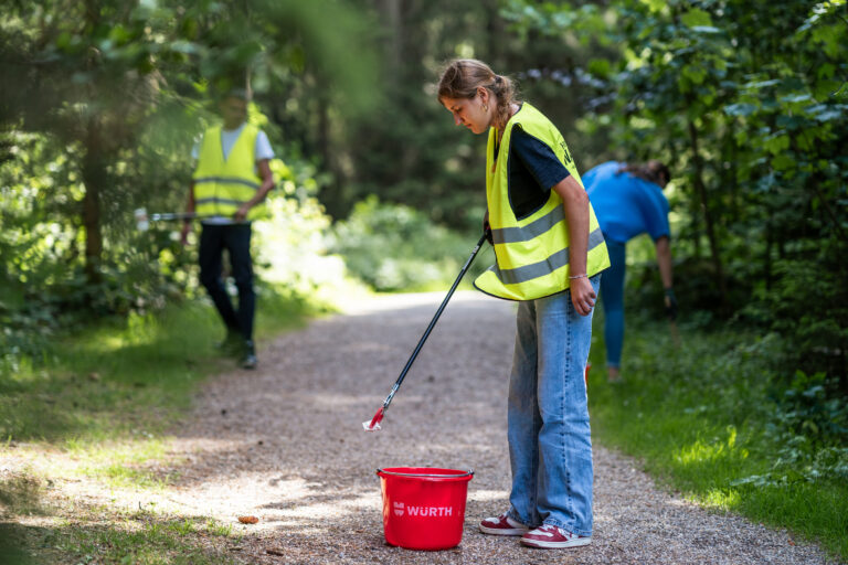Woman in a high-visibility vest uses a litter picker on a park path; a red Würth bucket sits nearby with green surroundings in the background.