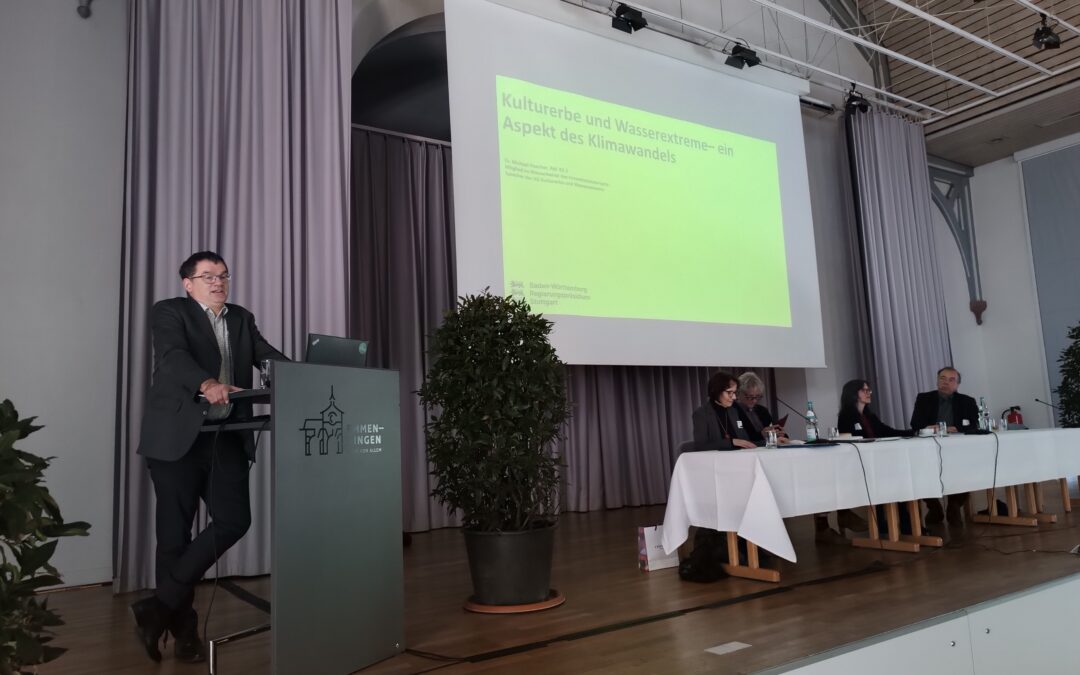 Man speaks at a podium on a stage during a conference, with a lime-green slide projected on screen and a panel of five people seated to the right, flanked by curtains and potted plants.