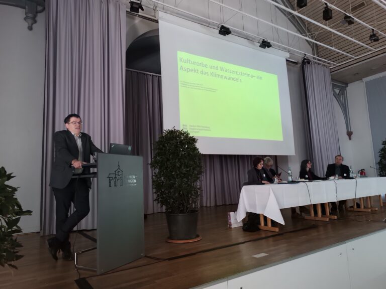 Man speaks at a podium on a stage during a conference, with a lime-green slide projected on screen and a panel of five people seated to the right, flanked by curtains and potted plants.