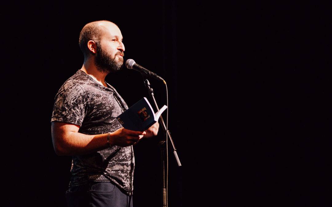 Bearded man in a patterned shirt reads from an open book into a microphone on a dark stage backdrop.