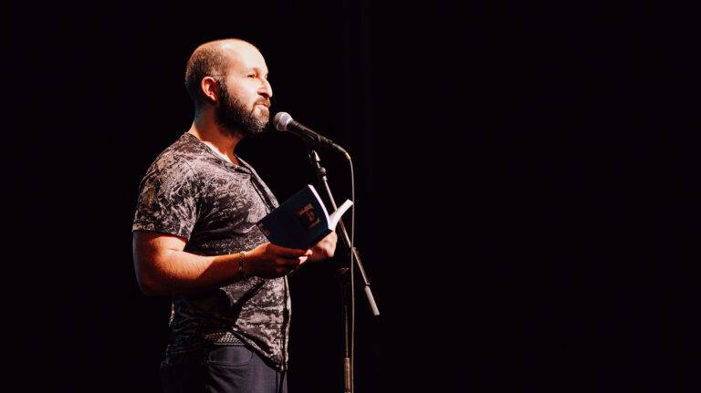Bearded man in a patterned shirt reads from an open book into a microphone on a dark stage backdrop.