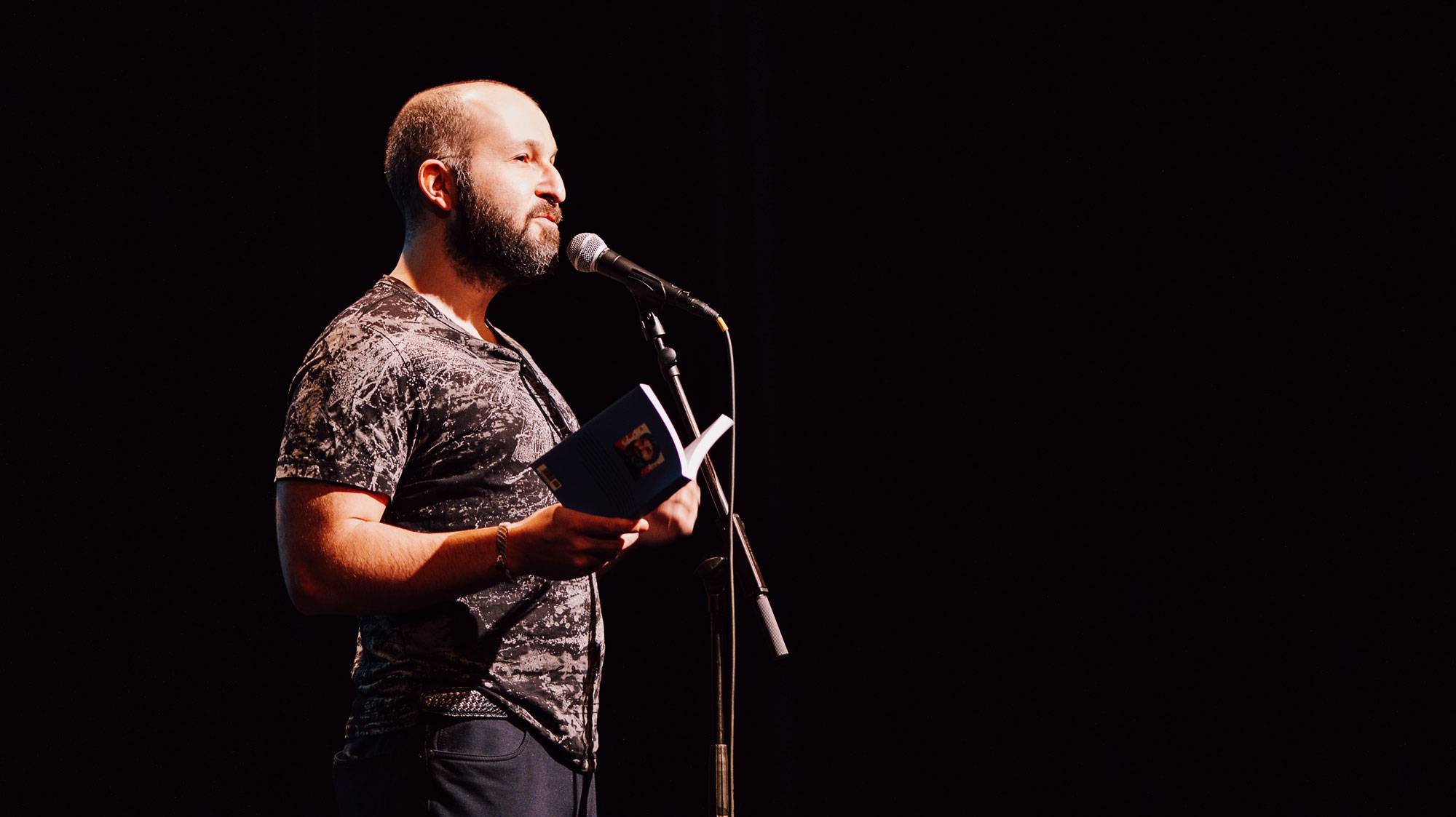 Bearded man in a patterned shirt reads from an open book into a microphone on a dark stage backdrop.
