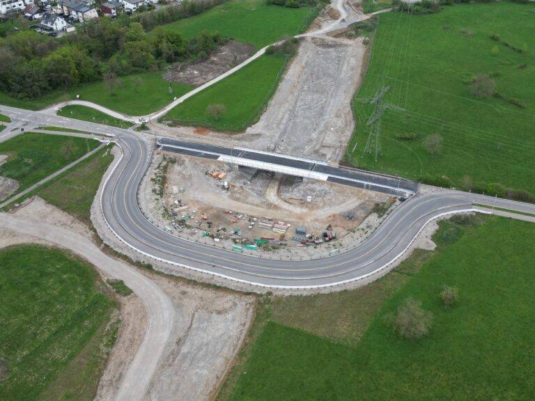 Aerial view of a curved highway under construction with an overpass and construction materials in a dirt worksite, surrounded by green fields and a nearby residential area.