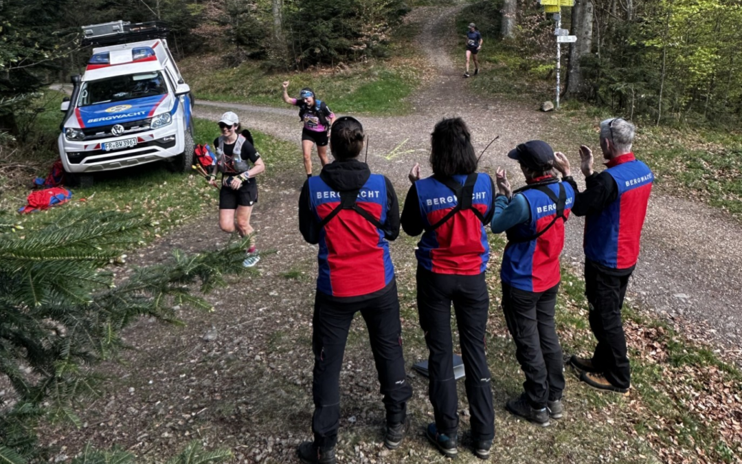 Bergwacht rescue team in red‑blue jackets watches a runner on a forest trail beside a white emergency SUV.