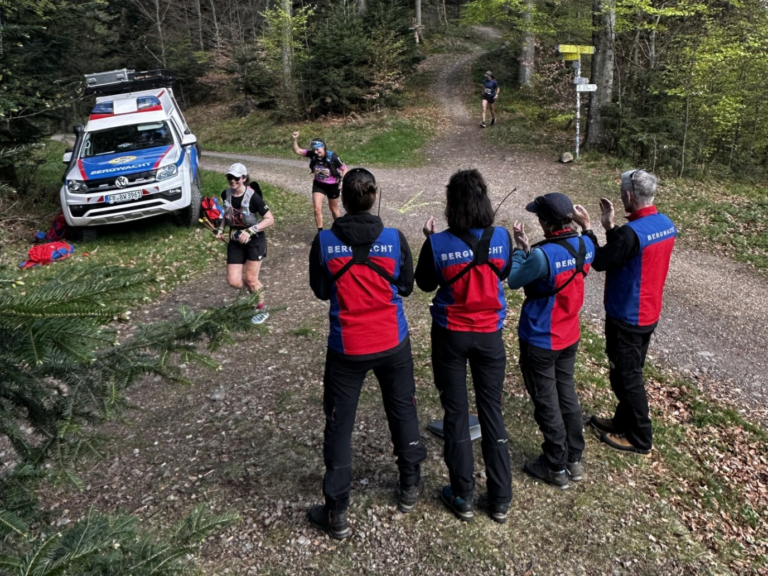 Bergwacht rescue team in red‑blue jackets watches a runner on a forest trail beside a white emergency SUV.