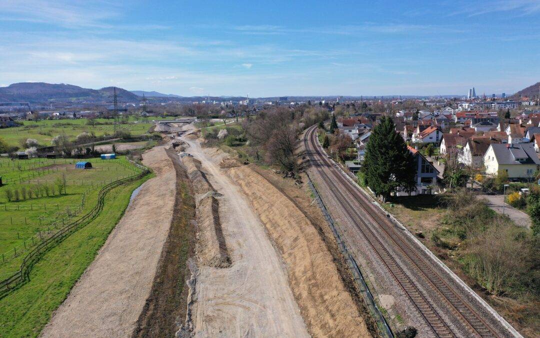 Aerial view of a construction site beside a railway line, with green fields and a town in the distance under a blue sky
