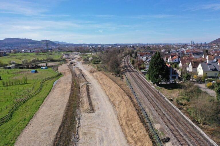 Aerial view of a construction site beside a railway line, with green fields and a town in the distance under a blue sky