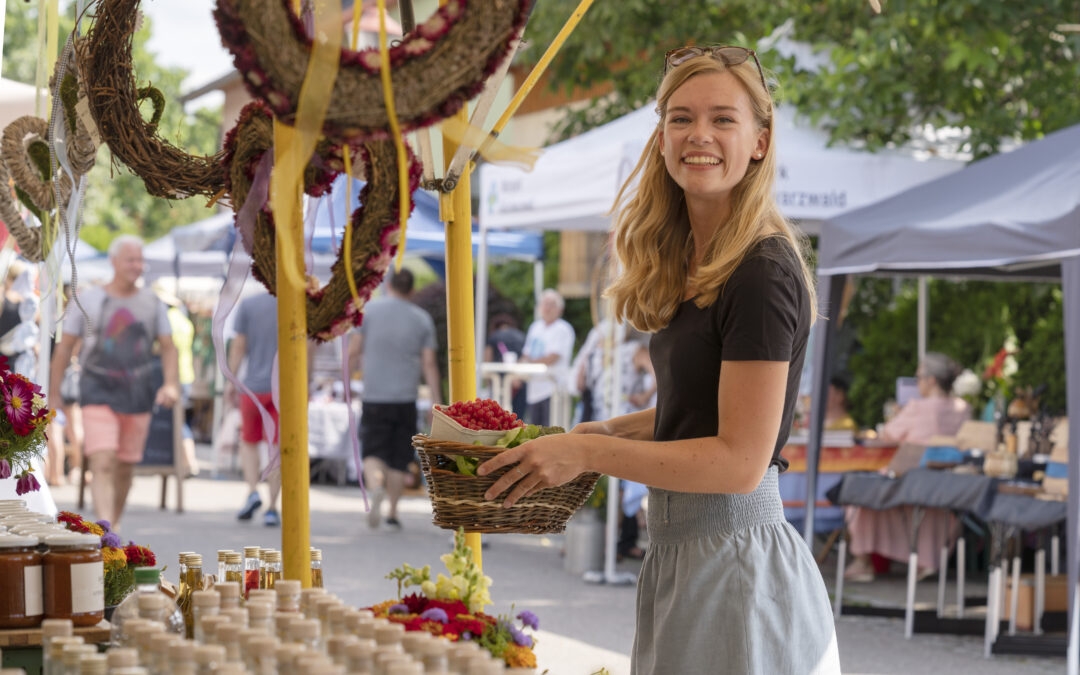 Smiling young woman at an outdoor market holds a wicker basket of berries beside a stall with jars and flowers in the foreground.