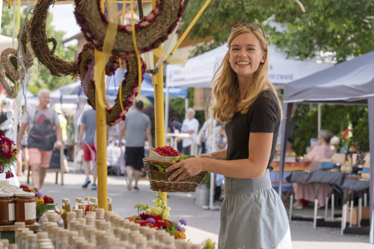 Smiling young woman at an outdoor market holds a wicker basket of berries beside a stall with jars and flowers in the foreground.