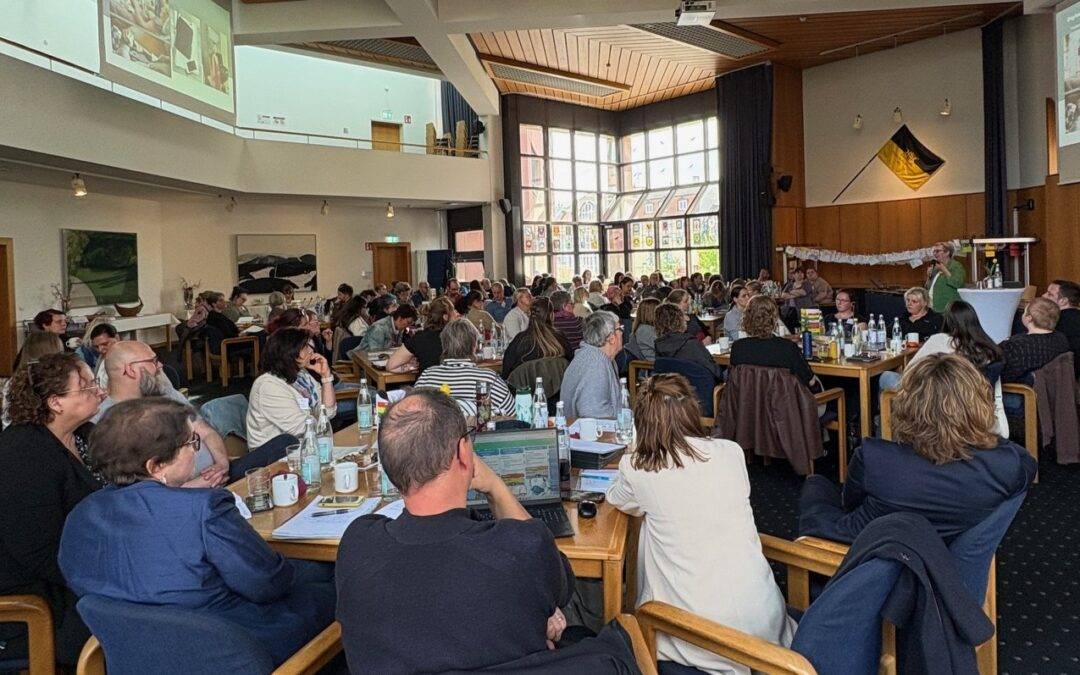 Large conference room filled with people seated at tables, a speaker at a podium on stage addressing the audience.