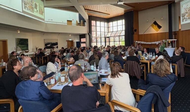 Large conference room filled with people seated at tables, a speaker at a podium on stage addressing the audience.