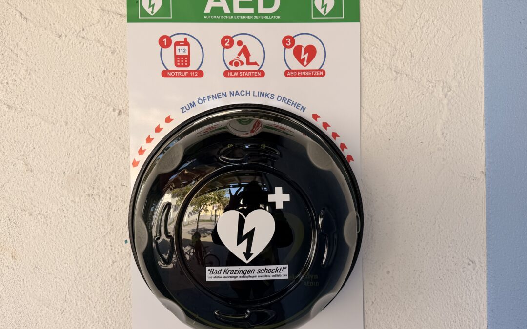 AED safety panel on a wall with three instructional icons (emergency number 112, start CPR, use AED); a black shiny helmet sits in front of it.