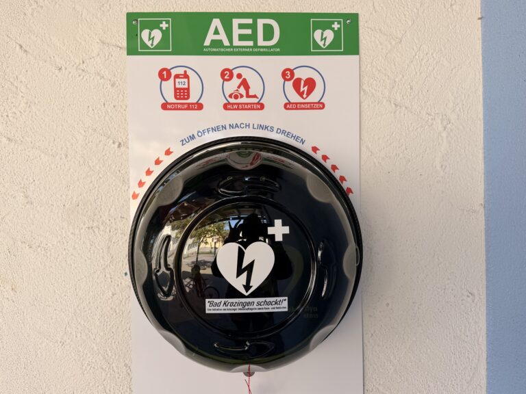AED safety panel on a wall with three instructional icons (emergency number 112, start CPR, use AED); a black shiny helmet sits in front of it.