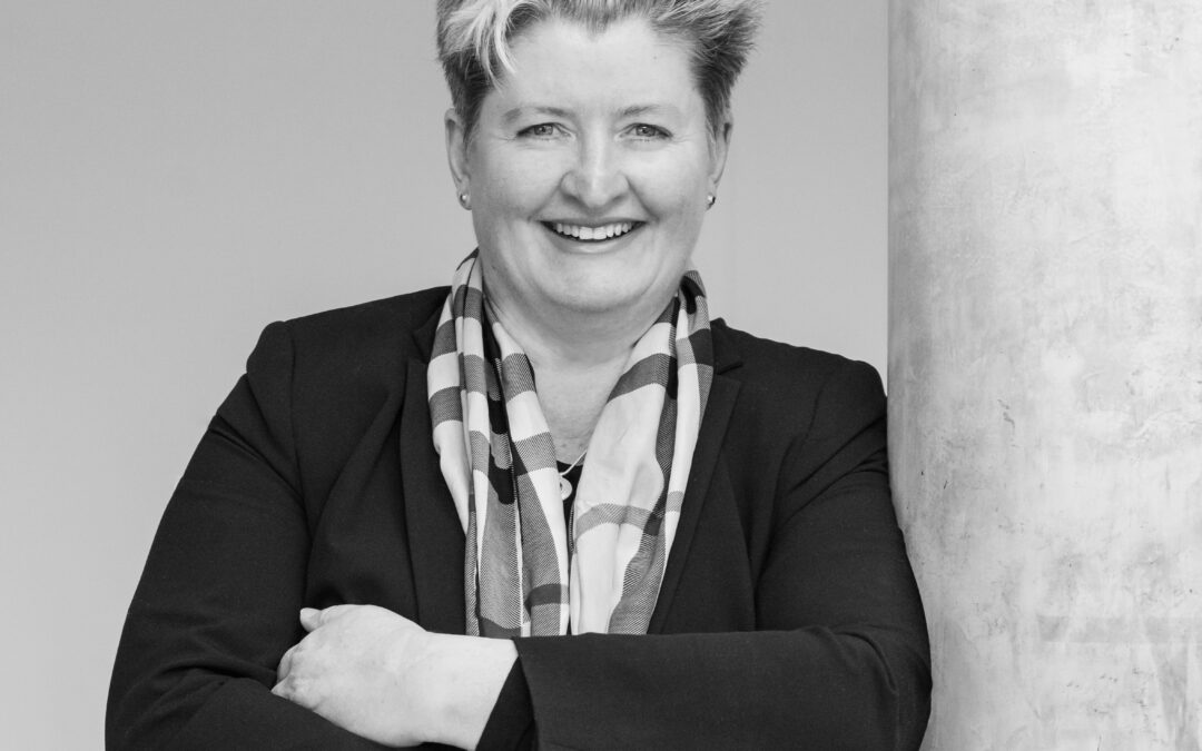 Smiling woman in a black blazer and patterned scarf with arms crossed beside a concrete column. But concise: "Smiling professional woman with arms crossed, standing beside a column."