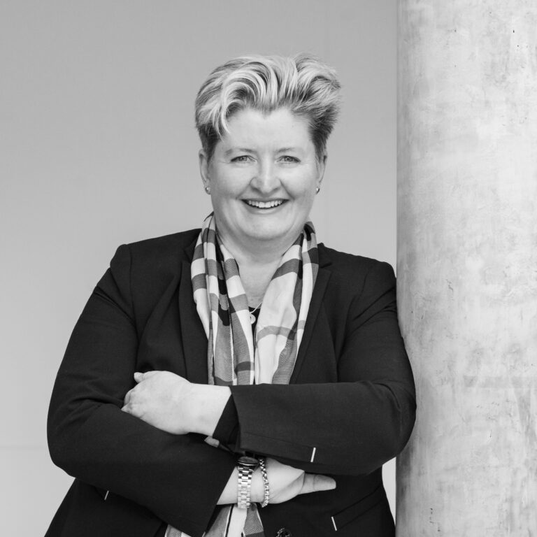 Smiling woman in a black blazer and patterned scarf with arms crossed beside a concrete column. But concise: "Smiling professional woman with arms crossed, standing beside a column."
